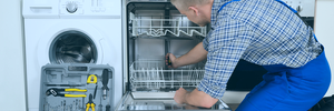 A white goods repair engineer in blue overalls inspecting a dishwasher in a customer's kitchen